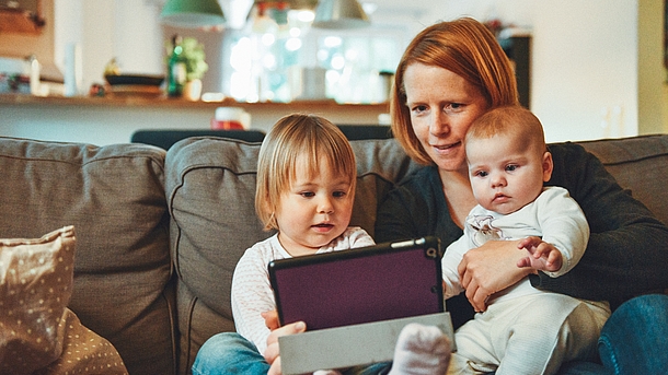 Frau sitzt mit mit zwei Kleinkindern auf dem Sofa und alle schauen auf ein Tablet.